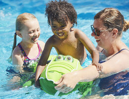 In a pool, a woman holds a floating toy for two smiling kids.