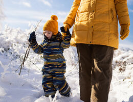 A young child in a snowsuit walks through snow with an adult in a parka.