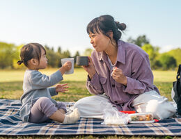 A woman and toddler on a picnic blanket, mugs raised.