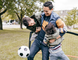 A man lifts two small boys as he kicks a soccer ball in a park.